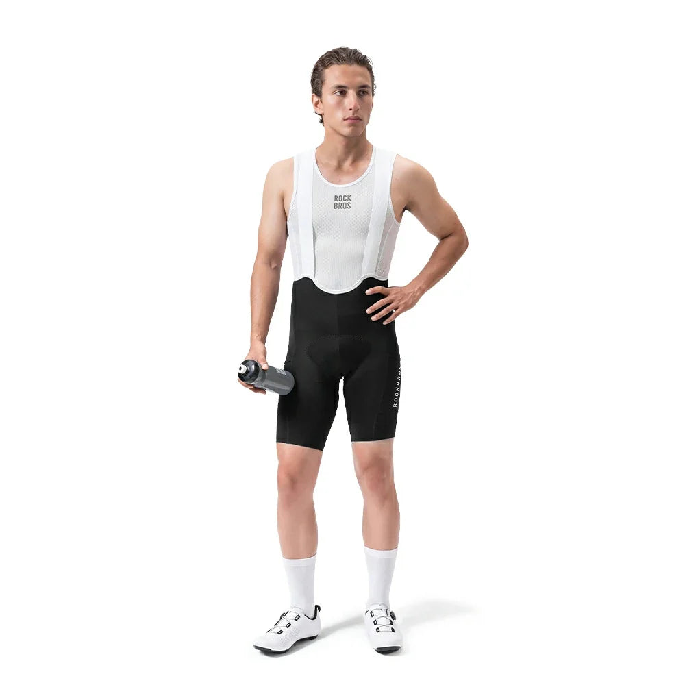 A male cyclist stands against a white studio background wearing a white bib shirt with black suspenders and black cycling shorts. He is holding a gray water bottle and wearing white socks and cycling shoes.
