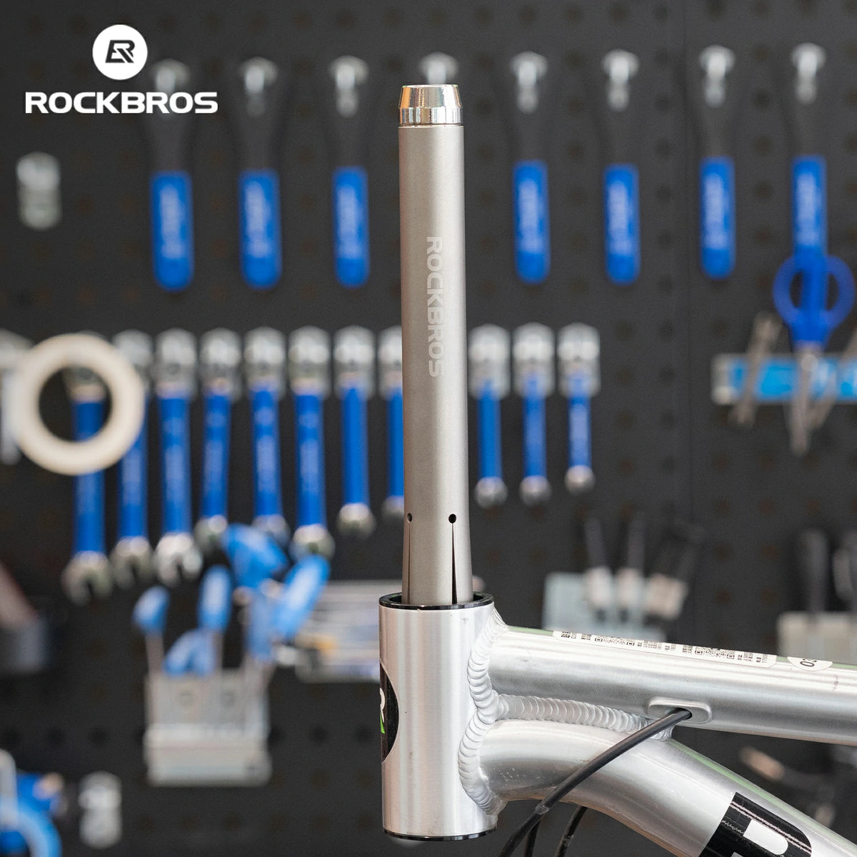 A silver Rockbros bicycle headset component mounted on a silver bicycle stem, with a blurred workshop background showing a pegboard with blue-handled tools and the Rockbros logo in the top left.