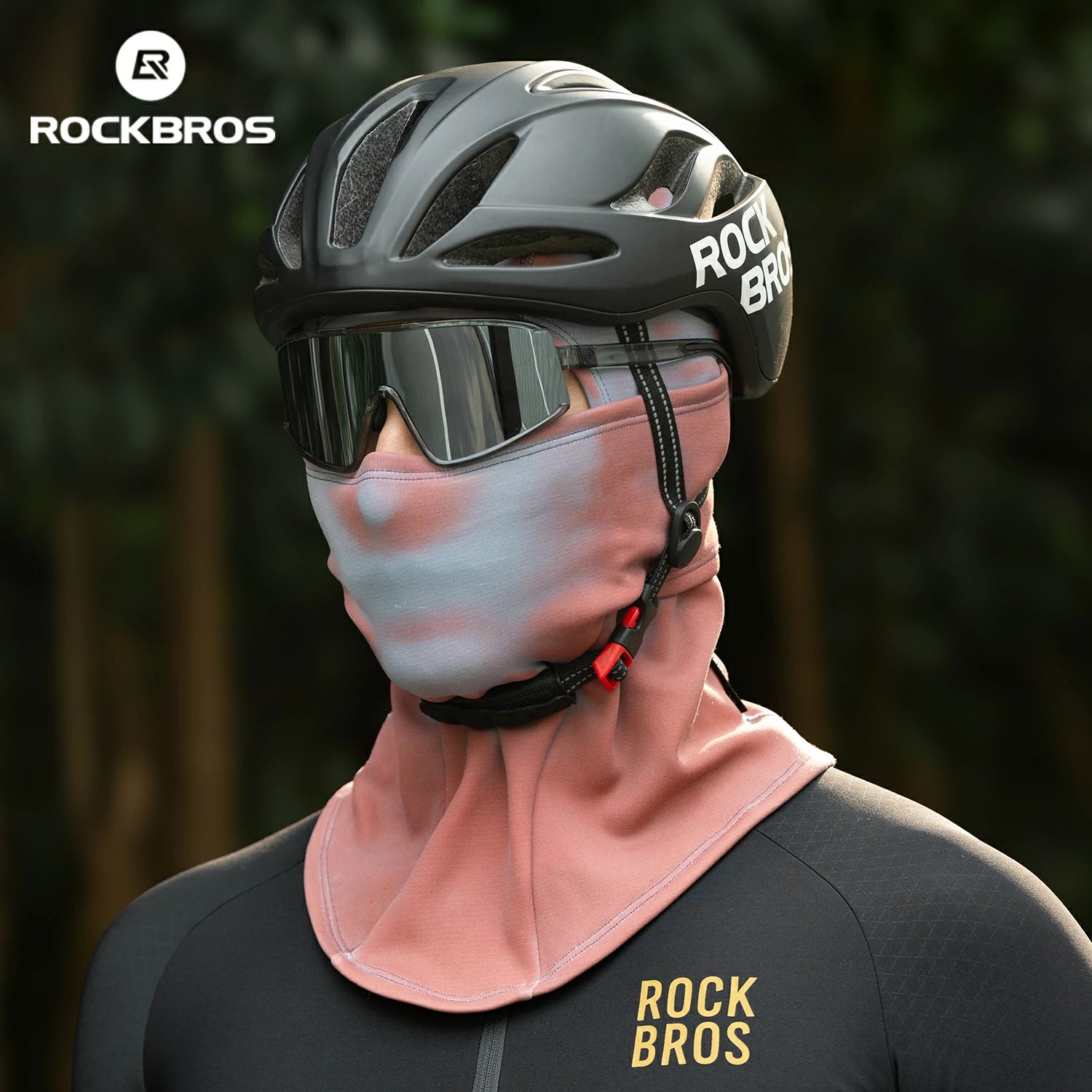 Person wearing a black helmet and pink face mask with 'RockBros' branding against a blurred natural background. #color_Brick red
