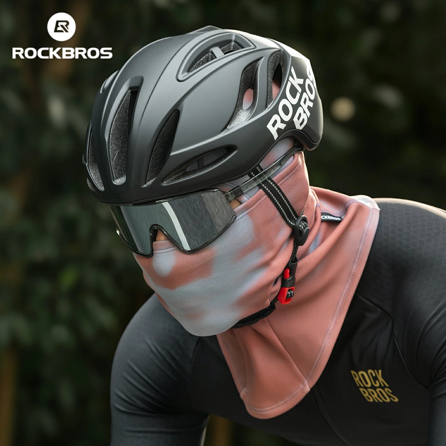 Person wearing a RockBros helmet and black cycling outfit with a blurred green background #color_Brick red