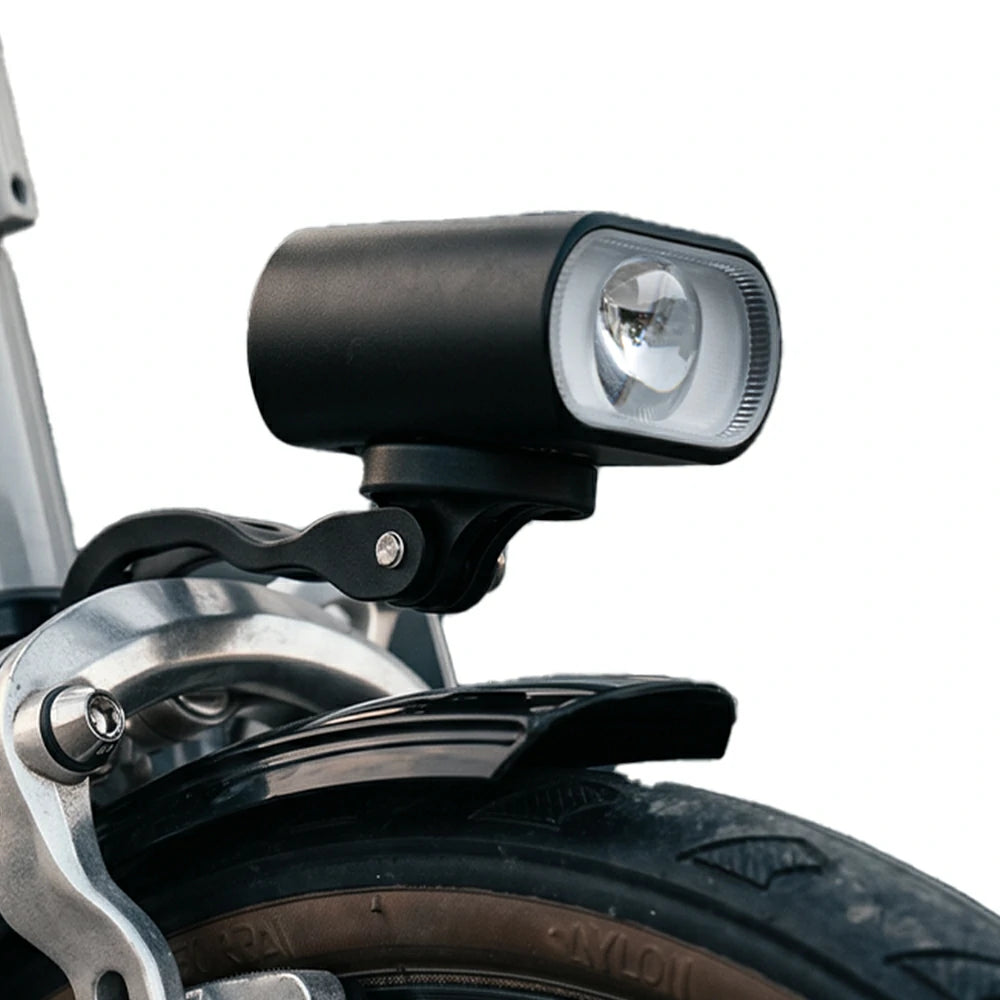 A close-up shot of a black bicycle headlight mounted on a silver bicycle frame above a tire, set against a plain white background.
