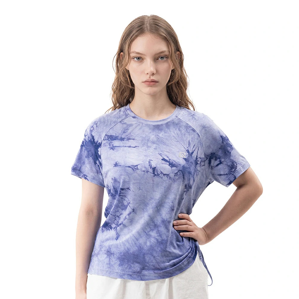 A woman modeling a short-sleeve crewneck t-shirt with a blue and purple tie-dye pattern, paired with white pants, against a solid white background.