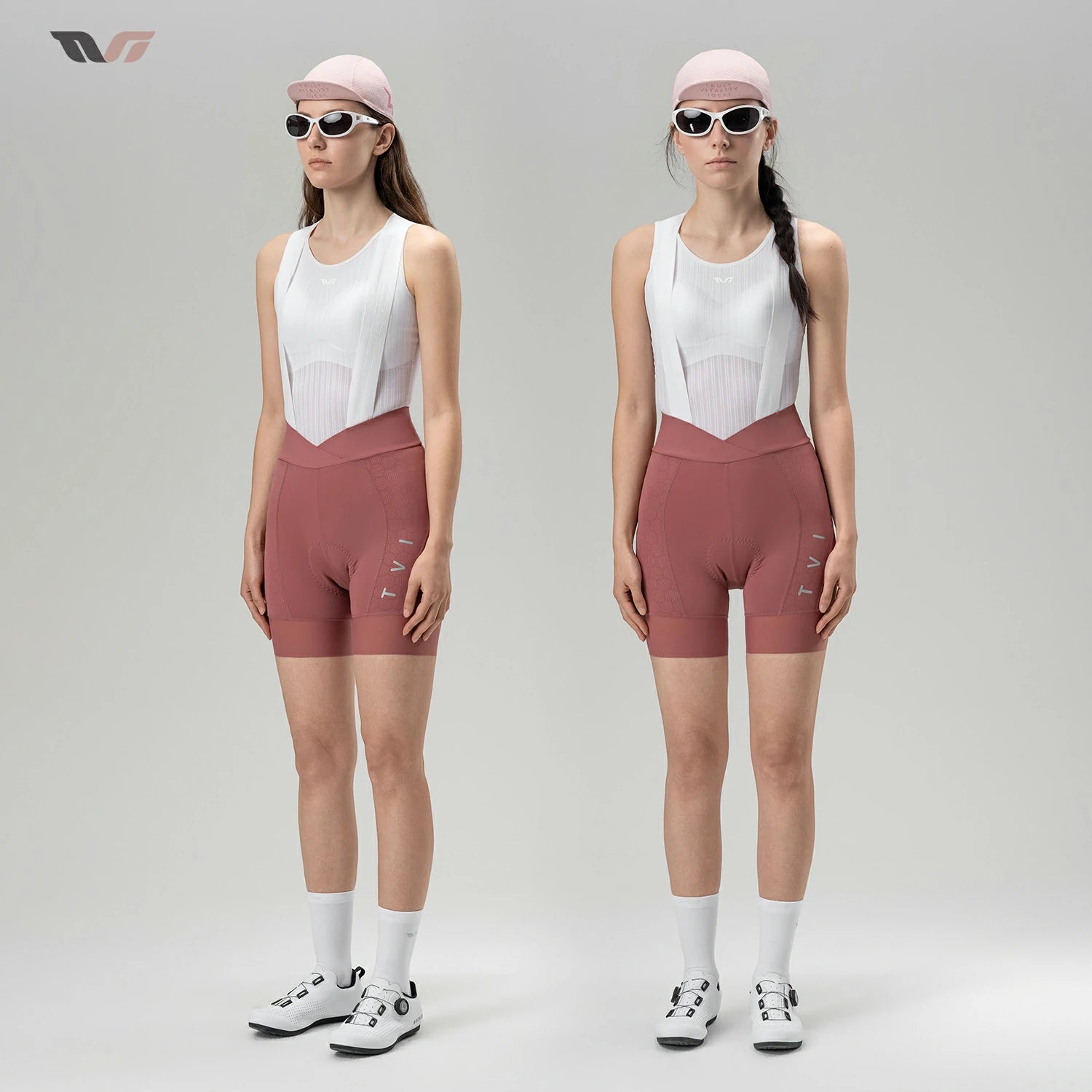 Two female models standing side by side in a studio, wearing matching athletic cycling outfits consisting of white bib shorts and dusty rose fitted shorts, white socks, and cycling shoes. Both wear light pink caps and dark sunglasses against a light gray background.