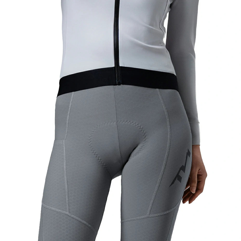 Close-up rear view of a person wearing gray cycling tights with a black waistband and a black logo on the upper thigh, paired with a white and gray long-sleeve top against a white background.