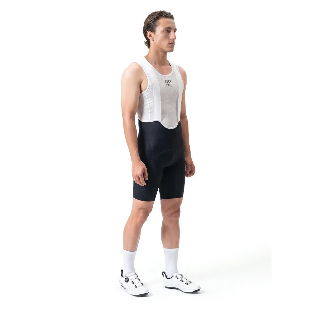 A male cyclist stands in profile wearing a white aerodynamic bib shorts, black cycling shorts, white socks, and white cycling shoes against a plain white studio background.