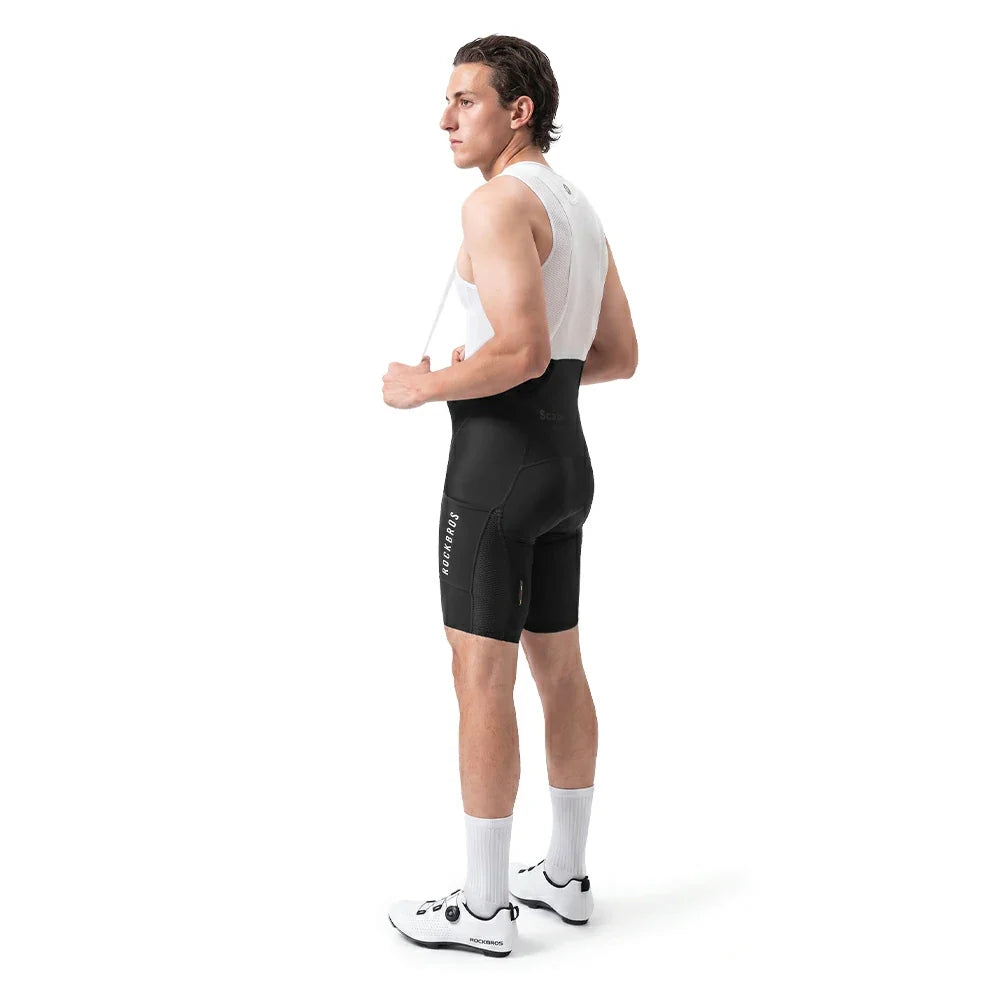 A man in a white cycling jersey and black cycling shorts stands in profile against a clean white background, wearing white cycling shoes and white socks.