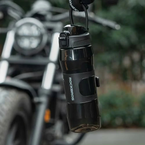 Black water bottle with 'Rockbros' branding hanging from a bike handlebar against a blurred natural background.