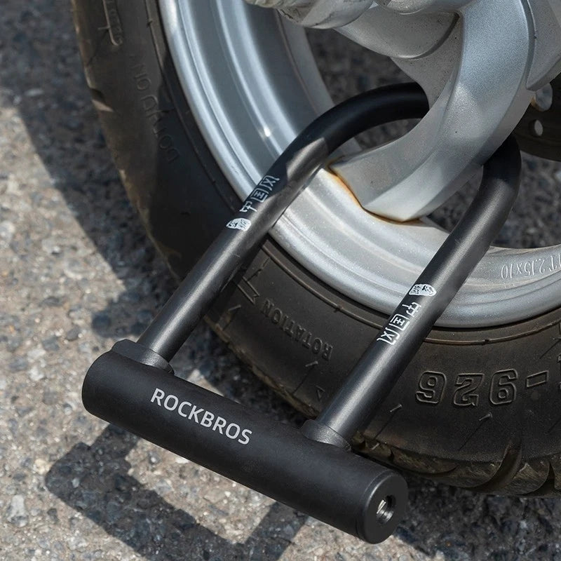 A close-up of a black U-lock bicycle wheel lock branded ROCKBROS, secured around a silver metal wheel rim on a paved ground.