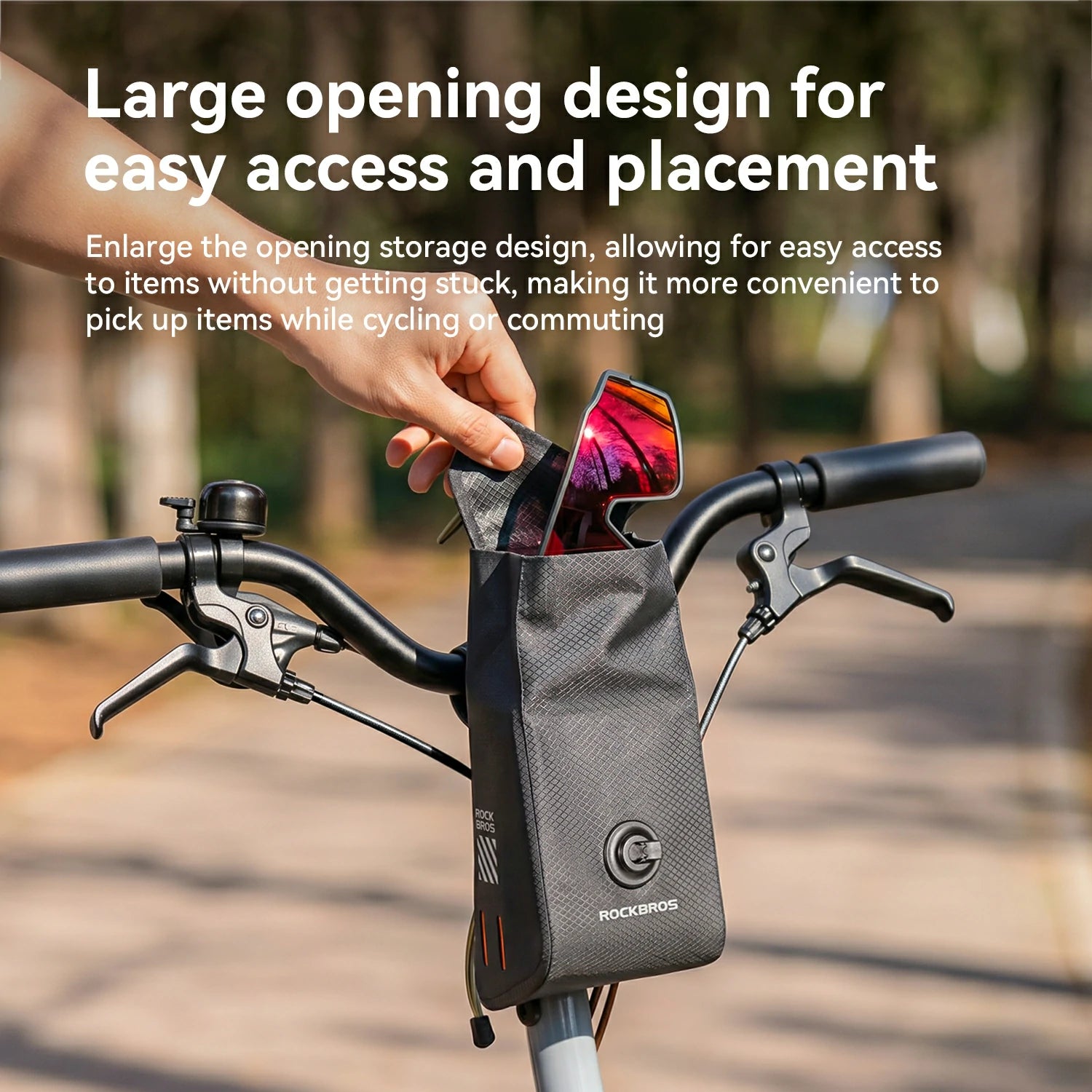 A black Rockbros bicycle handlebar bag mounted on a bike handlebar, with a hand reaching into the top opening to place or retrieve sunglasses. The background is a blurred outdoor path with trees.