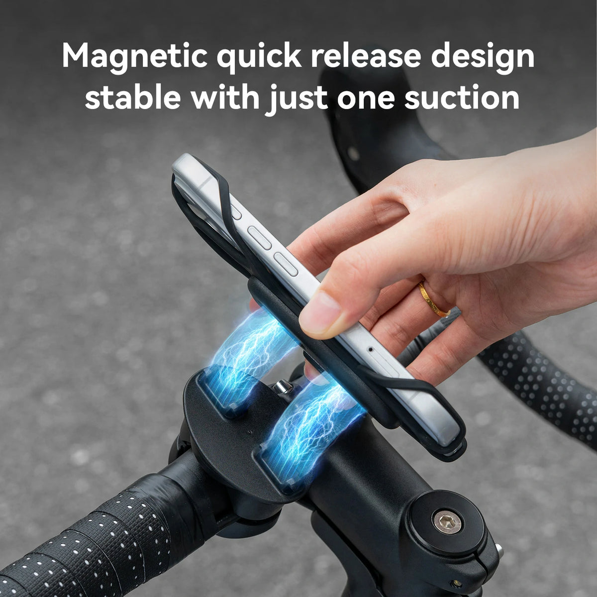 A hand holds a smartphone in a black magnetic bike handlebar mount attached to a black handlebar. Blue lightning graphics illustrate magnetic suction, with text reading 'Magnetic quick release design stable with just one suction' over a gray pavement background.