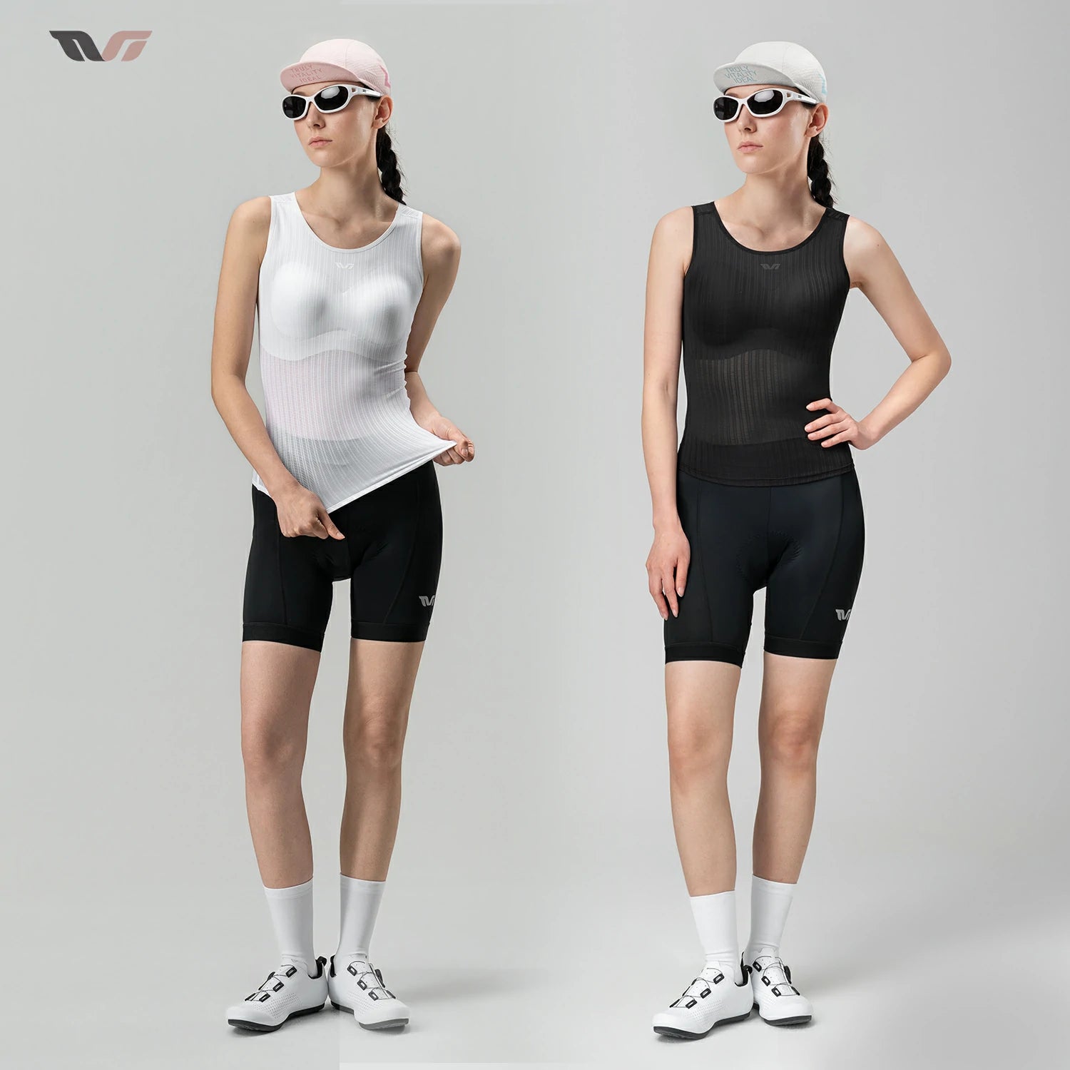 Two female models standing side by side in a studio, each wearing a sleeveless athletic compression top and matching fitted shorts. The left model wears a white top with black shorts, a pink cap, sunglasses, white socks, and white shoes.