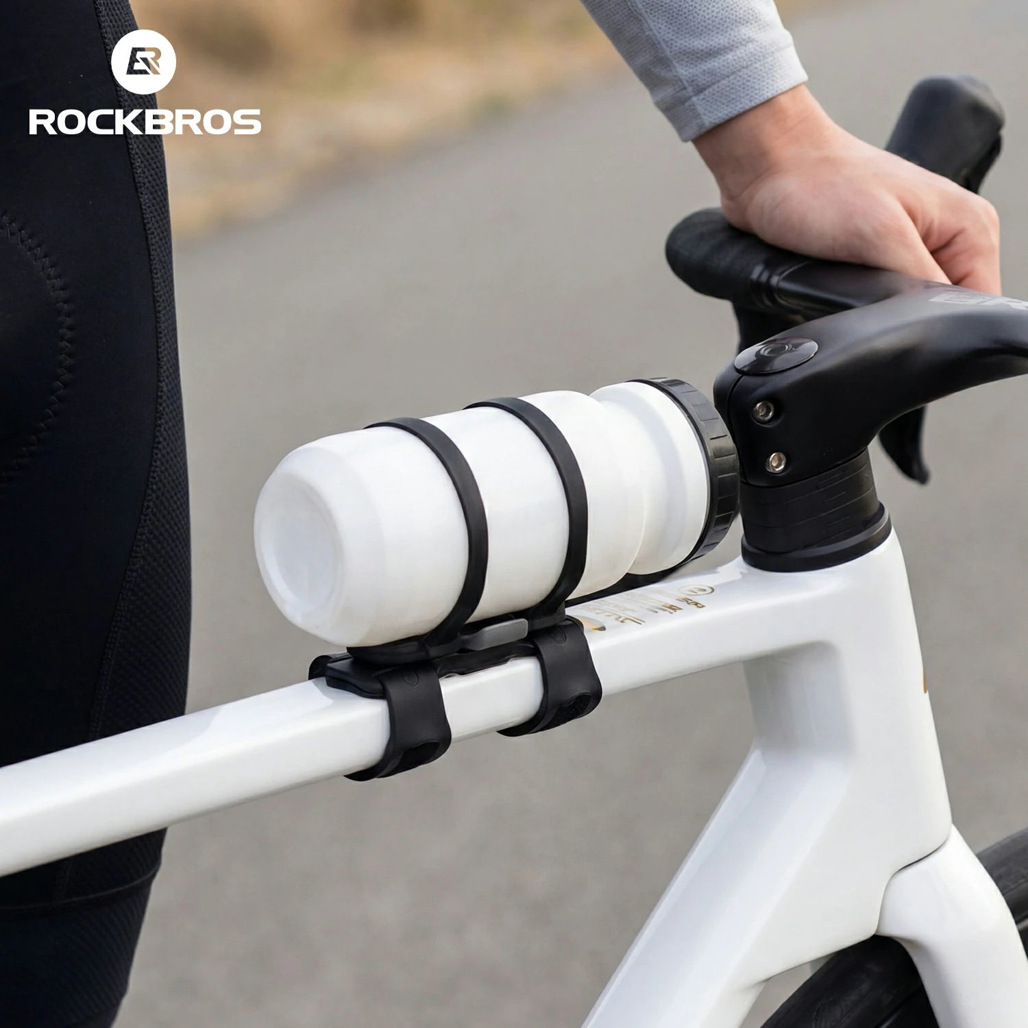 A close-up of a white water bottle mounted on a white bicycle frame with black straps, held by a rider whose hand and arm are visible, with the ROCKBROS logo in the top left corner.