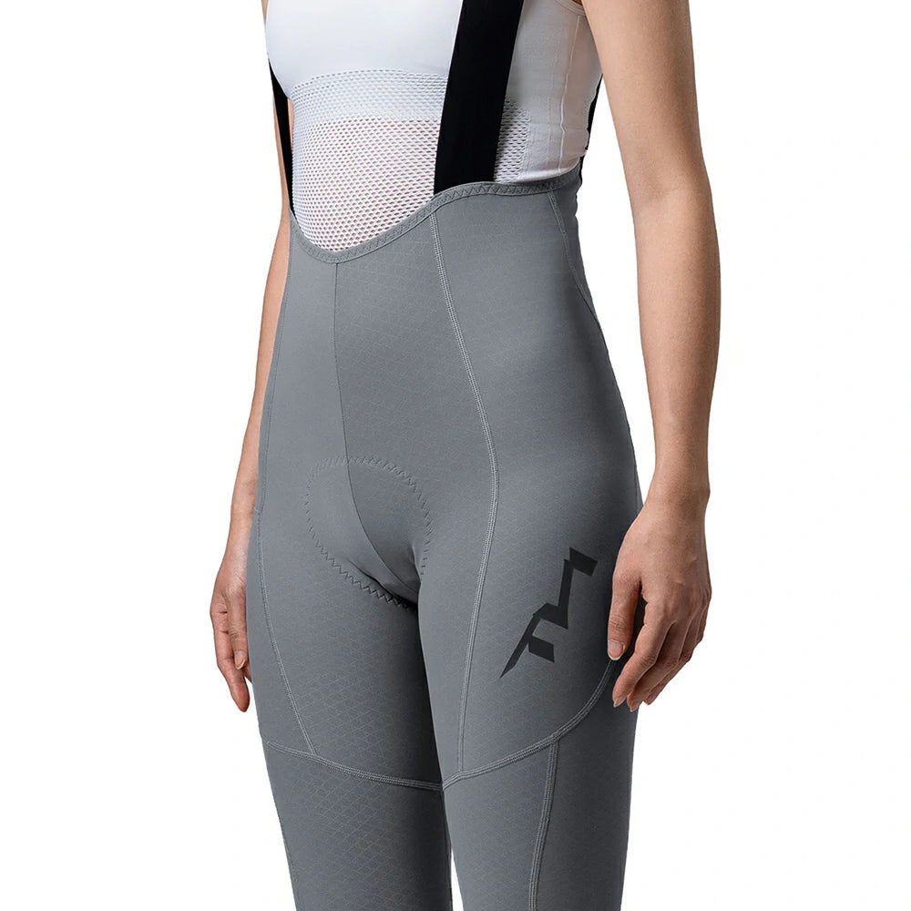 A close-up of a model wearing gray high-waisted cycling bib tights with black suspenders and a black logo on the thigh, set against a plain white background.