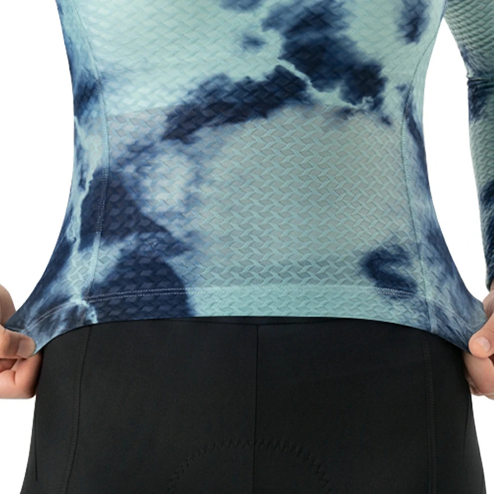 Close-up of a person wearing a long-sleeve athletic top with a blue and mint tie-dye pattern, stretching the fabric at the sides to show elasticity, paired with black cycling shorts.