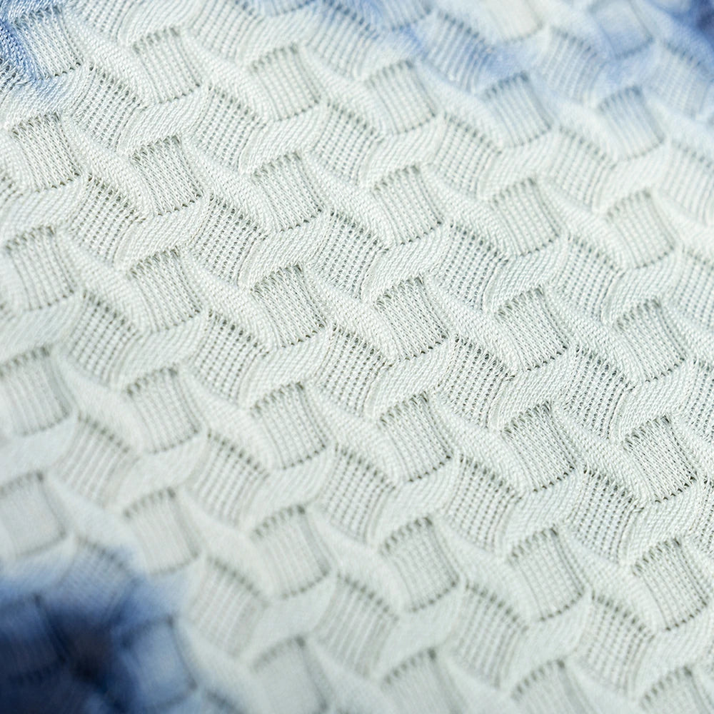 Close-up of a textured knit fabric with a repeating geometric diamond pattern in off-white and light blue tie-dye accents.